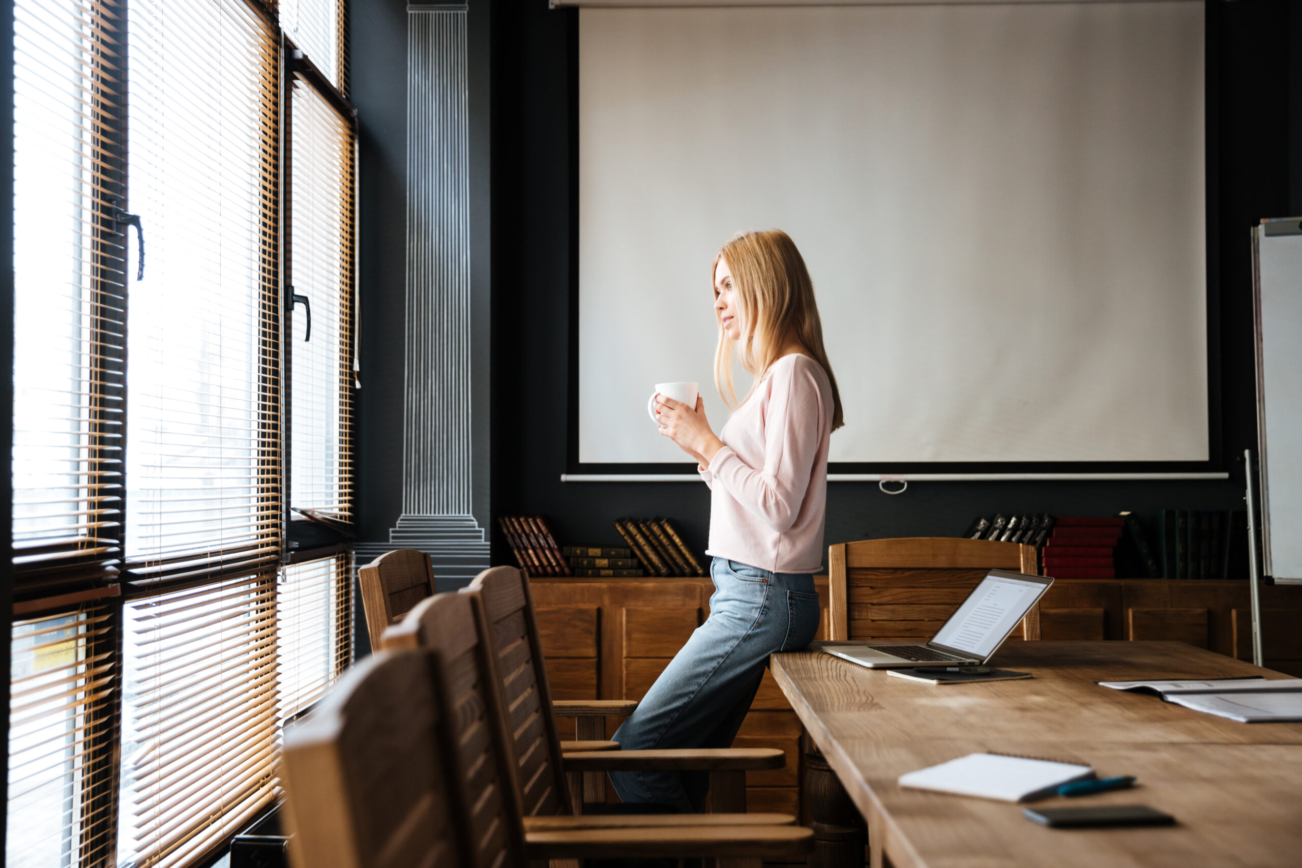 Young female employee standing in work with coffee in hands and laptop next to her